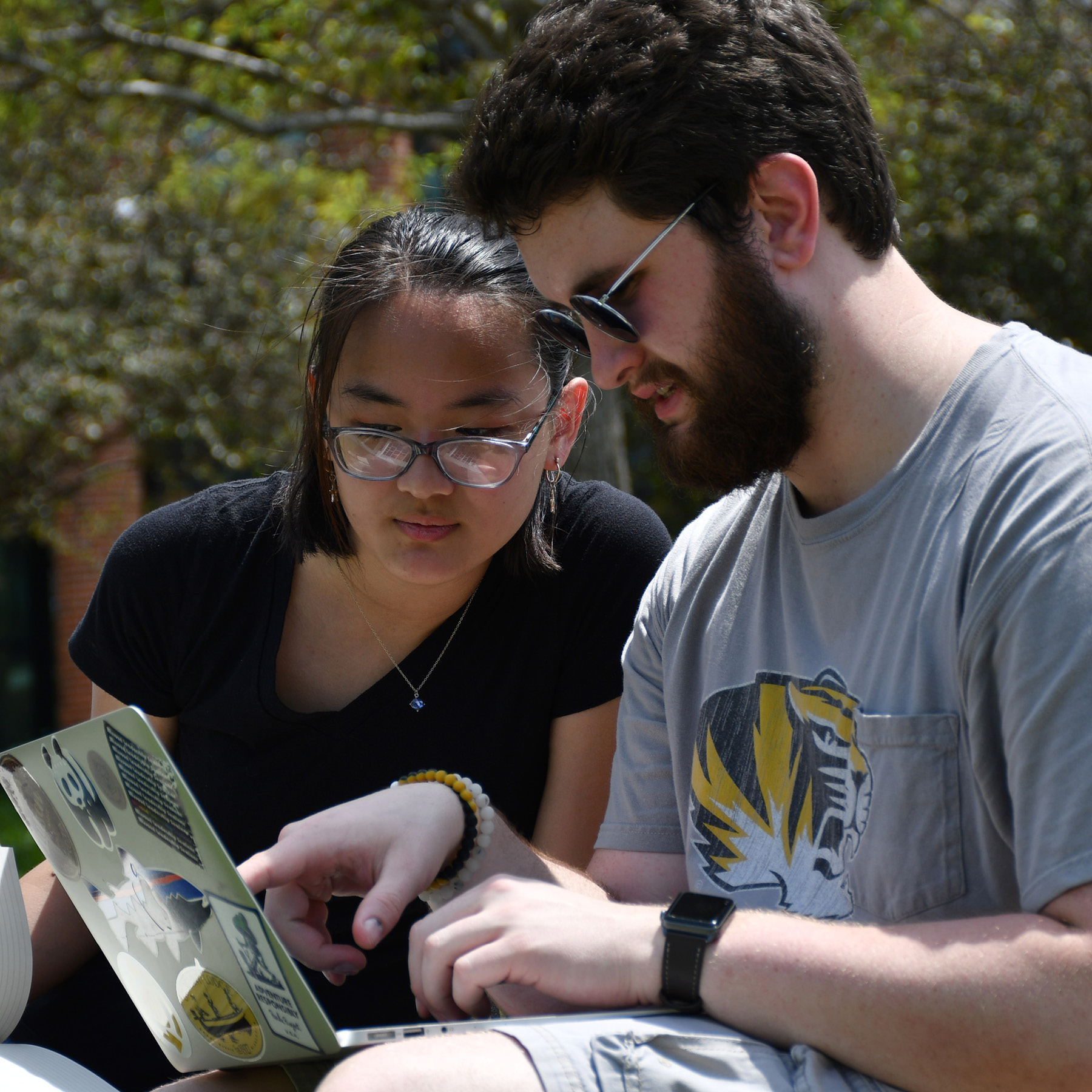 Two students sit outdoors looking at an assignment on a laptop.