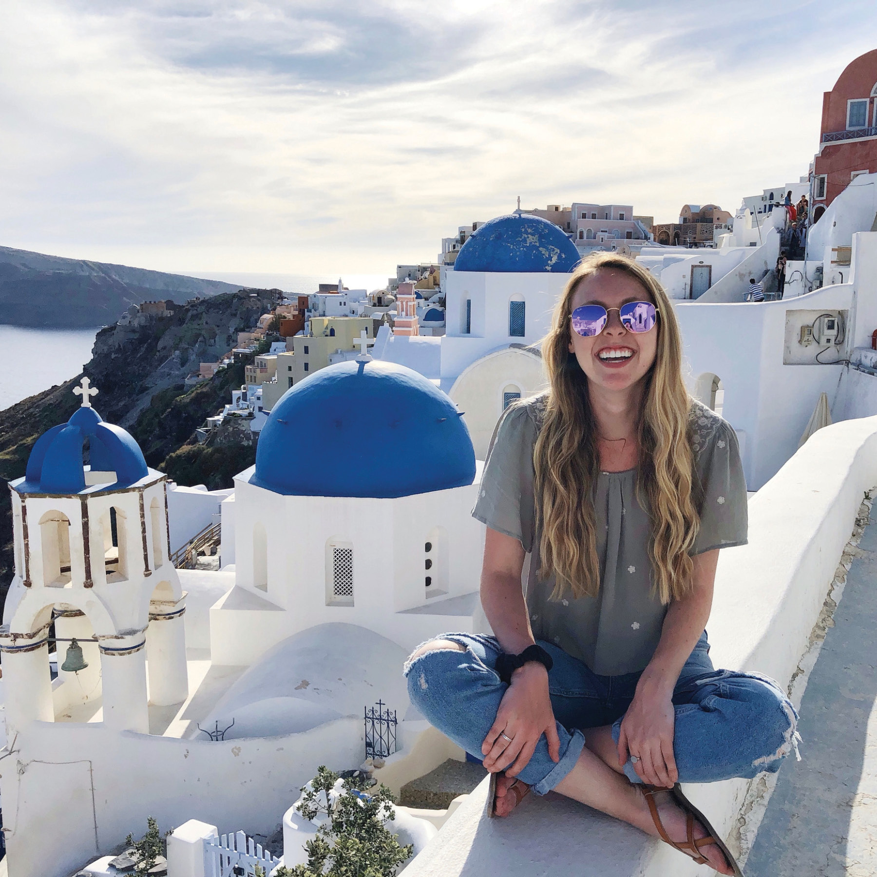 A smiling student sits on a wall with a beautiful view of the Greek island of Santorini behind her.