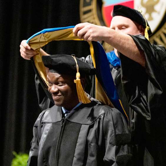 A student in cap and gown smiles as an honorary stole is placed on his shoulders at commencement